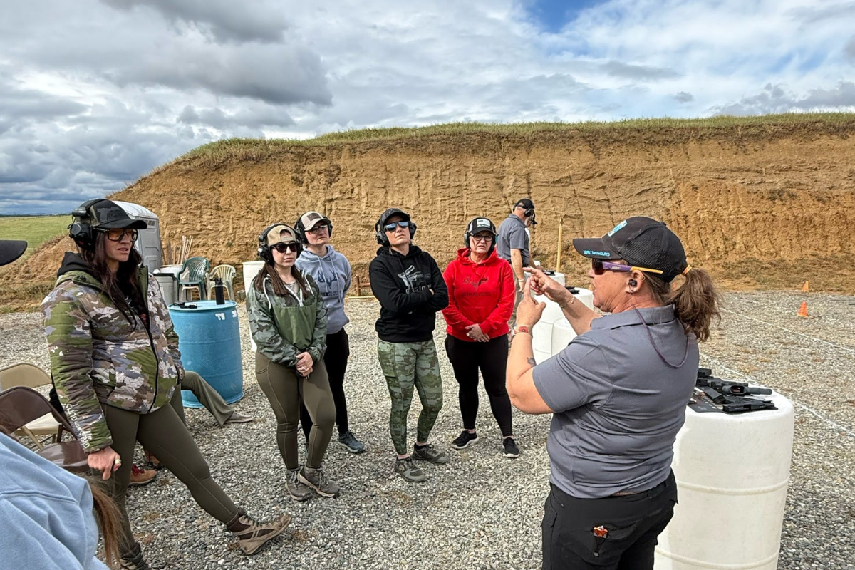 A firearms instructor demonstrates shooting techniques to a small group of participants at an outdoor shooting range. The group stands on a gravel range wearing eye and ear protection while listening to the instructor.